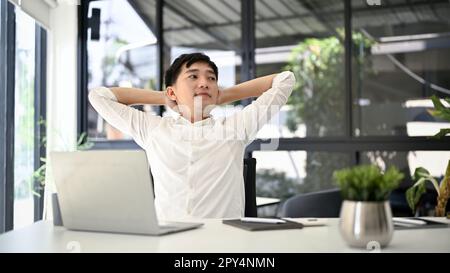 Un homme d'affaires asiatique détendu du millénaire s'est penché sur sa chaise de bureau, s'étirant et mettant ses bras derrière sa tête pour se détendre après avoir terminé son travail. Banque D'Images
