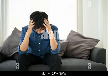 Un homme asiatique millénaire stressé et frustré est assis sur un canapé dans un salon, souffrant de ses factures de ménage et contrarié par sa vie et sa carrière. Banque D'Images