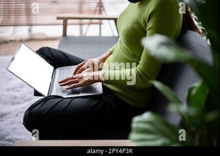 Image courte vue latérale d'une femme vêtue de vêtements décontractés en utilisant son ordinateur portable sur un canapé dans un salon. maquette d'écran blanc d'ordinateur portable Banque D'Images