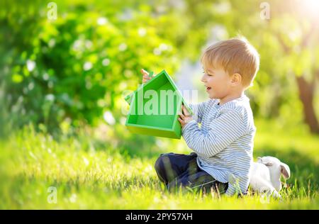 Enfant souriant assis sur la prairie de printemps avec le modèle de la maison verte dans ses mains. Banque D'Images