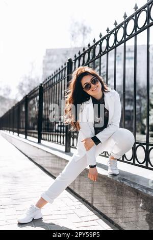 Belle femme brune étonnante avec une longue coiffure ondulée au printemps élégant vêtements de ville marche le long de la rue. elle sourit et regarde la caméra Banque D'Images