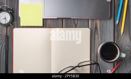 Pose plate de cahier vide ouvert, lunettes, crayons de couleur, stylo, horloge, tasse de café, ordinateur portable ouvert avec autocollant jaune. Banque D'Images