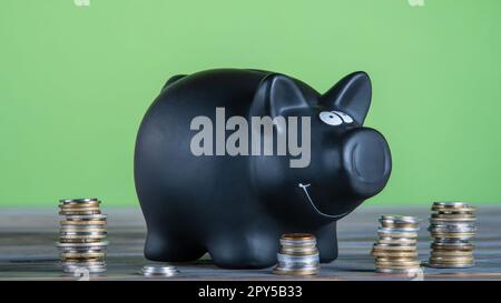 Tirelire en céramique noire souriante, caisse fixe près des piles de pièces de monnaie sur la table sur fond vert. Économies, argent. Banque D'Images