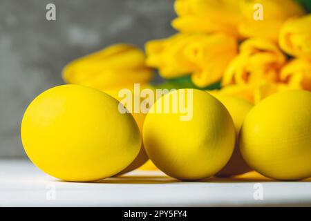 Bouquet de tulipes jaune vif et œufs de Pâques couchés sur une surface en bois blanc sur fond gris Banque D'Images