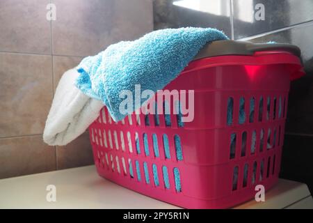 Serviettes dans le panier à linge. Les serviettes en éponge en coton bleu et rose sont jetées dans un panier en plastique rose. Entretien ménager. Stocker et séparer le linge avant du laver. Lumière provenant du haut de la fenêtre ouverte. Banque D'Images