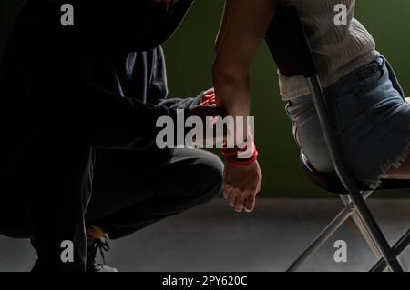 Criminel homme attaché les mains de femme avec la corde rouge. Prise d'otage femelle assise sur une chaise dans la chambre. Banque D'Images