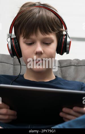 Sourire, brunette petit garçon dans un casque filaire regarder des films, écouter de la musique avec une tablette numérique dans les mains, s'asseoir sur un canapé Banque D'Images