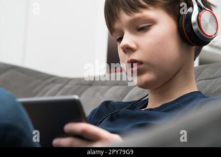 Calme concentré petit garçon dans le casque regarder des films et écouter de la musique avec la tablette numérique dans les mains, asseyez-vous sur le canapé Banque D'Images