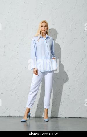 Femme en pantalon blanc, stilettos et blouse en coton bleu posant près du mur blanc Banque D'Images