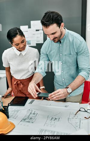 Leurs projets sont prêts à prendre vie. deux architectes utilisant un téléphone portable pour prendre des photos de plans dans un bureau. Banque D'Images