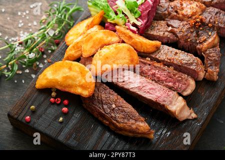 Steaks. Steak de viande grillée en tranches New York, Ribeye ou Chuck rouler avec garniture de salade et frites sur un plateau en marbre noir sur le vieux b bois Banque D'Images