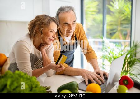 Les conjoints âgés enthousiastes utilisant un ordinateur portable et une carte de crédit dans l'intérieur de la cuisine, commander des articles d'épicerie ou la livraison de nourriture en ligne Banque D'Images