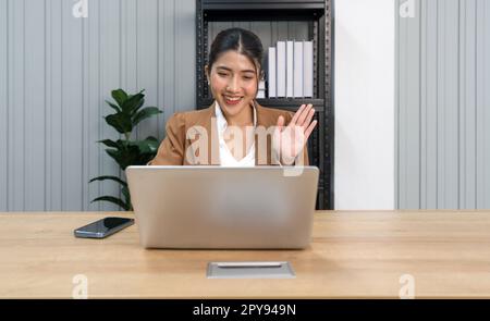 Jeune femme asiatique en costume marron, main levée sur un moniteur d'ordinateur portable, réunion en ligne avec un partenaire commercial. Concept « travail à domicile » Banque D'Images