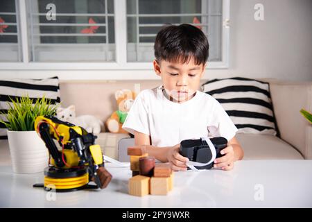 Joyeux petit garçon asiatique utilisant la télécommande jouant le bras de machine robotique pour ramasser le bloc de bois Banque D'Images