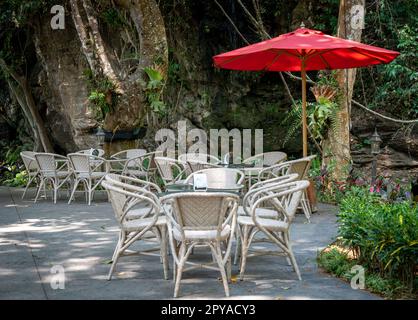 Un café en plein air, avec plusieurs tables et chaises disposées autour d'un parapluie rouge vif, sur fond d'arbres verts luxuriants Banque D'Images