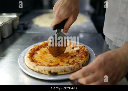 Chef cuisinier qui coupe une pizza fraîchement cuite avec un couteau rond sur une planche de bois Banque D'Images