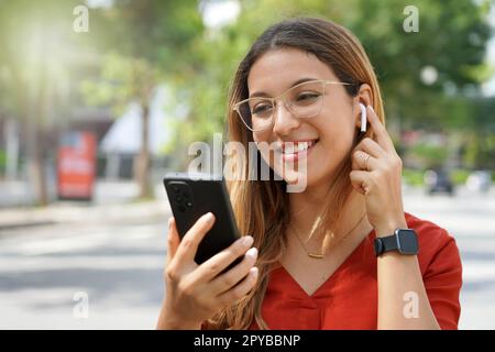 Gros plan sur les lunettes de protection, une fille allume des écouteurs sans fil pour écouter de la musique en choisissant une liste de lecture sur son smartphone en extérieur le matin Banque D'Images