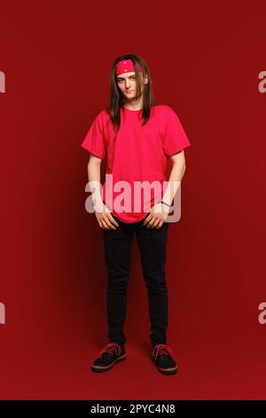 Portrait intérieur d'un homme hipster positif beau portant une bande rouge sur la tête, debout contre le mur rouge Banque D'Images