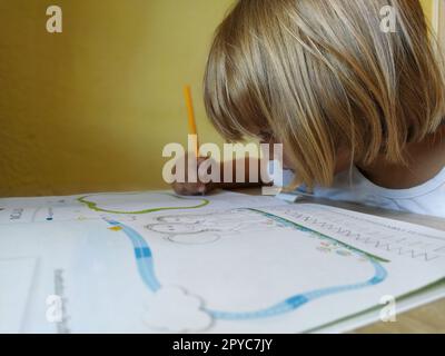 Une fille de 6-7 ans lit un livre ou un manuel et sourit. L'enfant a les cheveux blonds, la peau foncée et des lunettes sur son visage. L'enfant est assis au bureau. mur jaune en arrière-plan Banque D'Images