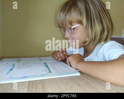 Une fille de 6-7 ans lit un livre ou un manuel et sourit. L'enfant a les cheveux blonds, la peau foncée et des lunettes sur son visage. L'enfant est assis au bureau. mur jaune en arrière-plan Banque D'Images