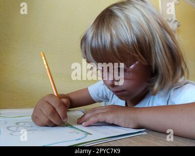 Une fille de 6-7 ans lit un livre ou un manuel et sourit. L'enfant a les cheveux blonds, la peau foncée et des lunettes sur son visage. L'enfant est assis au bureau. mur jaune en arrière-plan Banque D'Images