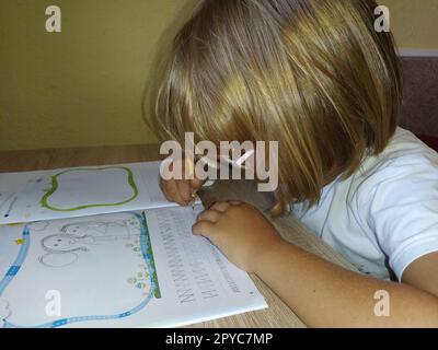 Une fille de 6-7 ans lit un livre ou un manuel et sourit. L'enfant a les cheveux blonds, la peau foncée et des lunettes sur son visage. L'enfant est assis au bureau. mur jaune en arrière-plan Banque D'Images