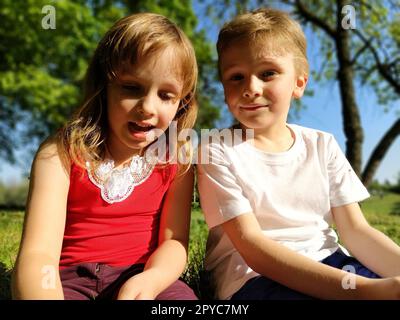 Deux enfants sont assis dans un pré et parlent. Une fille de 6 ans et un garçon de 7 ans jouent dehors dans un parc de la ville. Un garçon dans un T-shirt blanc, une fille dans un T-shirt rouge Banque D'Images