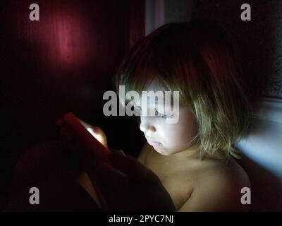 Mignon petite fille joue avec une tablette. Un enfant de 7 ans avec un visage sérieux et les cheveux blonds regarde dans un gadget. Espace de copie. Fille dans la chambre. Mur sombre et clair. À la maison la nuit Banque D'Images