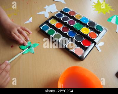 Bureau avec outils de dessin. Ensemble de peintures à l'aquarelle. Figurines de poisson peintes découpées en papier. Vue d'en haut. Leçon ou tâche créative. La main de l'enfant avec un pinceau peint une étoile Banque D'Images