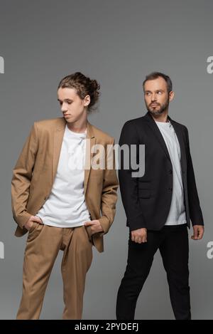 garçon adolescent bien habillé en costume beige debout avec les mains dans les poches à côté du père barbu isolé sur image grise Banque D'Images