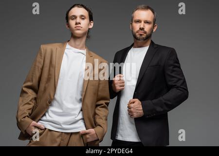 adolescent bien habillé en costume beige debout avec les mains dans les poches à côté du père barbu isolé sur gris, image de stock Banque D'Images