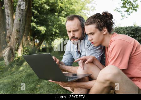 père et fils concentrés regardant l'ordinateur portable tout en étant assis ensemble sur la pelouse verte, image de stock Banque D'Images