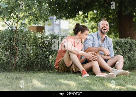 un père heureux rit tout en étant assis près d'un fils adolescent sur une pelouse verte dans le parc, image de stock Banque D'Images