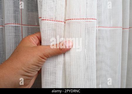 Rideaux blancs en coton épais ou en lin faits de fils fins. Intérieur d'appartement simple, compact et économique. Décoration de fenêtre dans la chambre. La main d'une femme fait un coup de tulle, montre la qualité. Banque D'Images