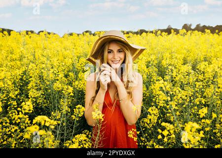 Portrait d'une jeune belle femme blonde, portant une robe rouge et un chapeau, pose au milieu d'un champ de fleurs de colza jaune en fleurs Banque D'Images