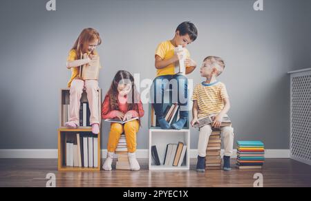 Groupe d'enfants assis sur les piles du livre et sur les étagères de la pièce et lisant le périodique. Banque D'Images