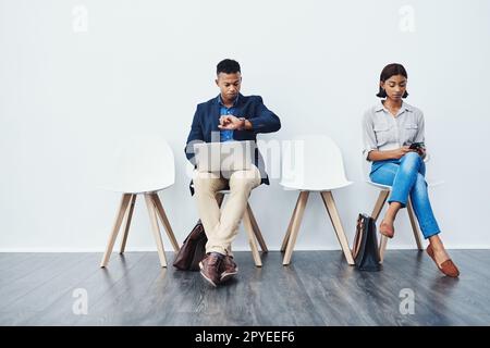 Nous attendions depuis un bon moment maintenant. Photo en pied de deux jeunes hommes d'affaires assis sur des chaises et utilisant leurs appareils technologiques sur un fond gris. Banque D'Images