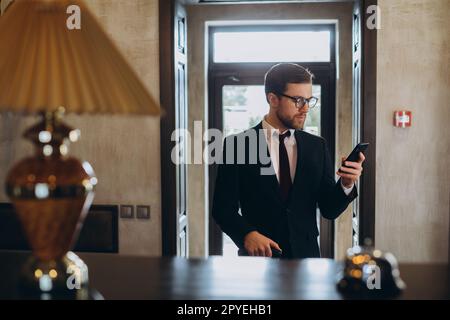 Businessman walking in hotel lobby avec valise et à l'aide de son téléphone intelligent. Homme d'affaires dans l'hôtel couloir avec téléphone et assurance. Banque D'Images