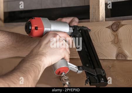 Gros plan les mains masculines recadrées utilisent une cloueuse pneumatique, un pistolet à clous agrafeuse pour planche et planche en bois. Vue première personne du constructeur Banque D'Images