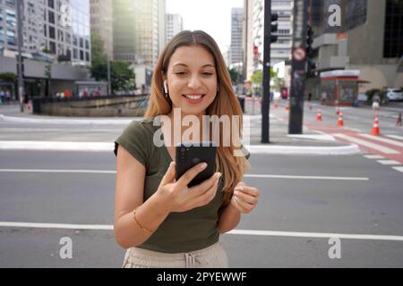 Une brésilienne excitée portant des écouteurs regarde son smartphone dans la rue Banque D'Images