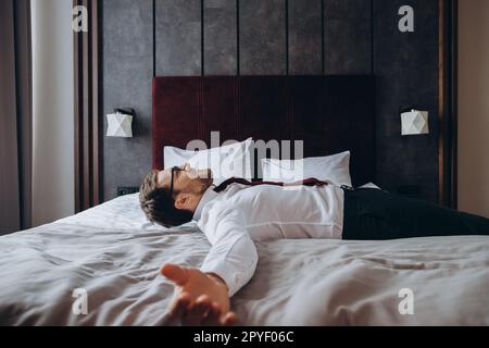 Homme d'affaires épuisé reposant sur le lit après un long vol d'air, décalage horaire. Homme en costume d'affaires allongé avec les bras étirés sur le matelas dans la chambre d'hôtel. Banque D'Images