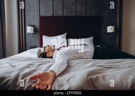 Homme d'affaires épuisé reposant sur le lit après un long vol d'air, décalage horaire. Homme en costume d'affaires allongé avec les bras étirés sur le matelas dans la chambre d'hôtel. Banque D'Images