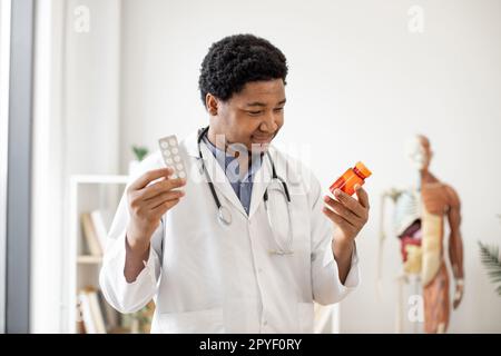 Homme multiethnique sympathique en laboratoire manteau scannant la bouteille de pilule tout en se tenant debout avec des comprimés dans le lieu de travail du médecin contre le fond anatomique modèle. Pharmacien efficace étudiant les médicaments d'ordonnance. Banque D'Images