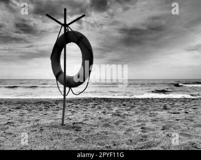 Bouée de sauvetage sur une plage de sable. Cercle orange sur un poteau pour sauver les personnes noyées dans la mer. Point de sauvetage sur le rivage. Ciel avec nuages et mer en arrière-plan. Photo noir et blanc avec contraste élevé. Banque D'Images