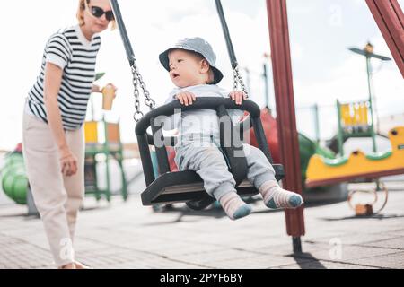 Mère poussant son bébé garçon enfant sur une balançoire sur le terrain de jeu à l'extérieur. Banque D'Images