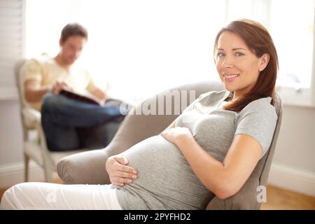 Créer une atmosphère détendue - grossesse. Portrait d'une belle femme enceinte tenant son estomac pendant que son mari est assis à l'arrière-plan. Banque D'Images