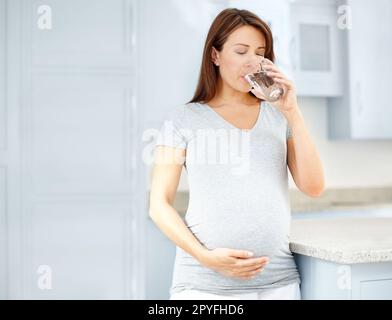 Prévention des complications. Une jeune femme enceinte boit un verre d'eau dans sa cuisine tout en tenant son ventre. Banque D'Images