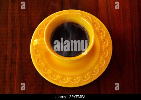 Goiania, Goias, Brésil – 01 mai 2023 : une tasse et une soucoupe jaunes remplies de café chaud à la vapeur sur une table en bois. Banque D'Images