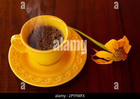 Goias, Brésil – 01 mai 2023 : une tasse jaune remplie de café chaud à la vapeur, avec une soucoupe et un tournesol, sur une table en bois. Banque D'Images