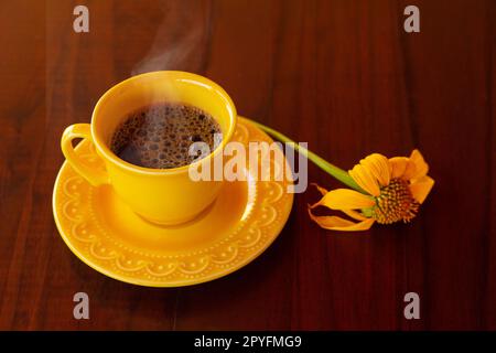 Goias, Brésil – 01 mai 2023 : une tasse jaune remplie de café chaud à la vapeur, avec une soucoupe et un tournesol, sur une table en bois. Banque D'Images